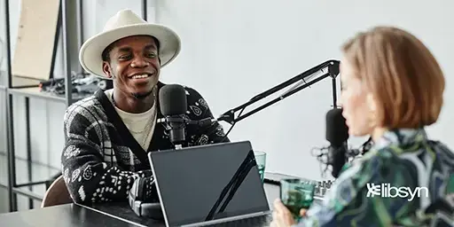 A white, female podcaster with bobbed hair is interviewing a smiling Black man with a lovely white hat and casual grey shirt