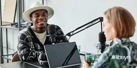 image - Official Libsyn Blog Two podcast hosts in a studio: a smiling man wearing a hat and a patterned sweater speaks into a microphone while a woman sits across from him, with a laptop and recording equipment on the table.